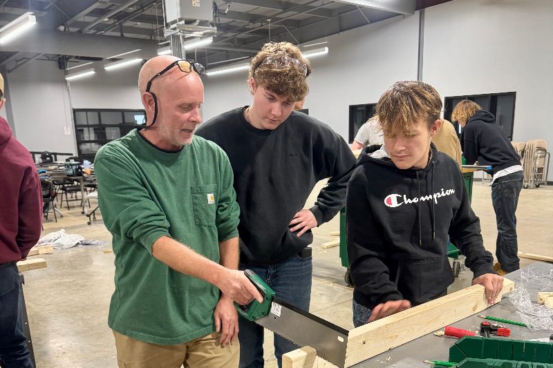 An instructor demonstrating a hand saw to two students at a workbench in the training area.